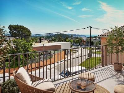 Terraza soleada con sillón de mimbre y mesa pequeña con vistas a un pueblo costero y al mar bajo un cielo azul.
