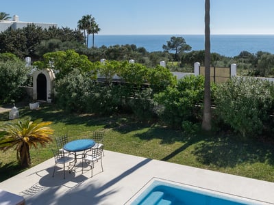 A sparkling blue swimming pool and patio set overlooking a lush garden and the Mediterranean Sea under a clear sky.