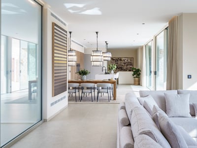 Amplio salón moderno con sofá gris claro, mesa de comedor de madera y lámparas de diseño junto a grandes ventanales.