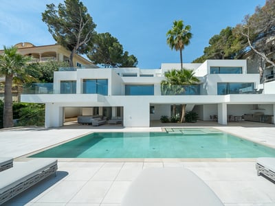 Modern white villa with a large turquoise pool, glass balconies, and palm trees under a clear blue sky.