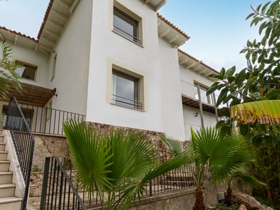 Fachada de casa blanca con base de piedra, escaleras exteriores y palmeras bajo un cielo despejado.