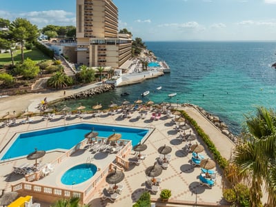 Aerial view of a swimming pool area with sunbeds and umbrellas overlooking a clear turquoise bay and rocky coastline.