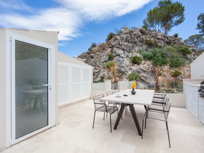 Terraza blanca moderna con mesa de comedor, sillas y zona de barbacoa, frente a una montaña rocosa.