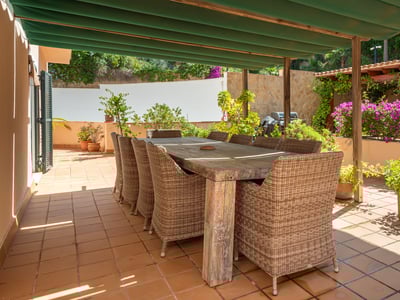 Gran mesa de comedor de madera con sillas de mimbre en una terraza soleada de terracota bajo un toldo verde.
