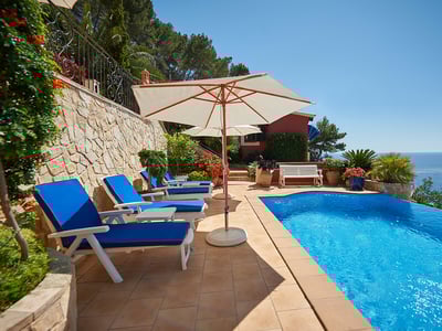 Piscina infinita en terraza de terracota con tumbonas azules y sombrillas blancas frente al mar bajo un cielo despejado.