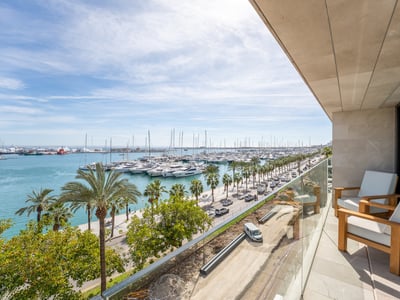 Modern balcony with wooden armchairs and glass railing overlooking a marina filled with yachts and palm trees.
