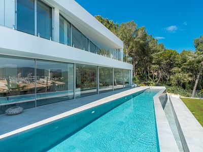 A sleek modern white villa with a long turquoise lap pool, floor-to-ceiling glass windows, and lush green trees.