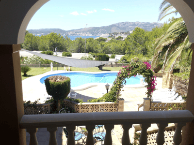 Vy genom en valvgång över en njurformad pool, solterrass och avlägsen havsutsikt omgiven av grönska.