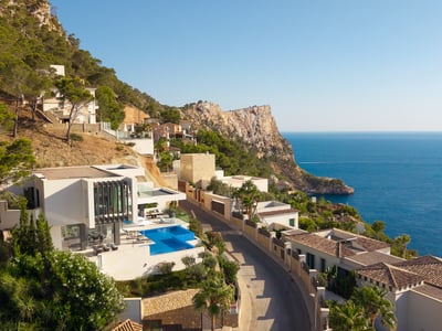 Aerial view of a white modern villa with an infinity pool on a cliffside overlooking the blue Mediterranean sea.