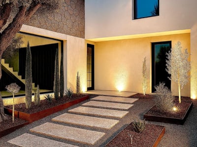 Camino de piedras hacia una casa moderna al atardecer con un olivo y luces cálidas integradas en el suelo.