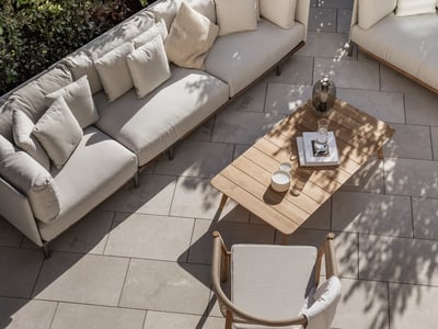 Draufsicht auf eine luxuriöse Terrasse mit beigen Sofas, Holztisch und Steinplatten im gefleckten Sonnenlicht.