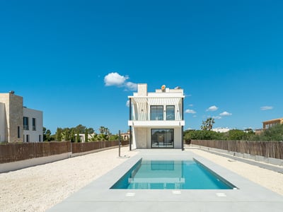 A rectangular blue swimming pool leading to a modern white two-story villa under a clear blue sky.