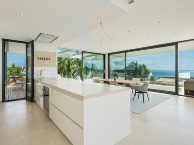 Cuisine blanche moderne avec grand îlot, salle à manger et baies vitrées du sol au plafond donnant sur l'océan.