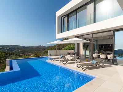 Luxury infinity pool and white modern villa with mountain views and designer loungers under white umbrellas.