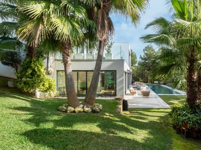 Grönskande trädgård med palmer som leder till en modern vit villa och en lång pool under en blå himmel.