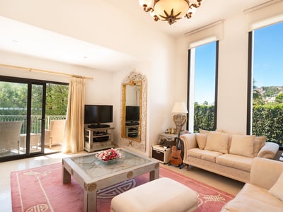 Salón luminoso con sofás crema, alfombra rosa, grandes ventanales verticales y puerta corredera hacia un balcón.