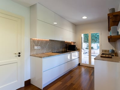 Cocina moderna de color blanco con encimeras de mármol, suelos de madera y puertas de cristal que dan a una terraza.
