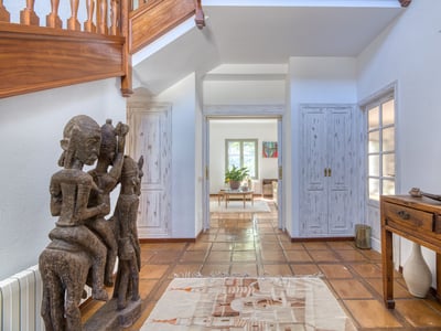 Amplio pasillo con suelos de terracota, una gran estatua tribal de madera y una escalera de madera hacia arriba.