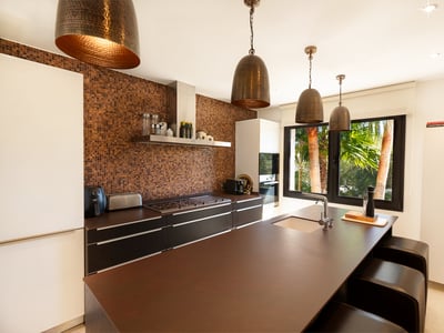 Cocina moderna con isla central marrón, salpicadero de mosaico cobrizo y lámparas colgantes de metal martillado.