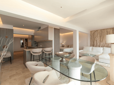 Modernes Wohnzimmer mit Glastisch, cremefarbenem Sofa und integrierter Küche in neutralen Tönen und Stein.