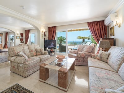 Bright living room with floral sofas, marble coffee table, and large glass doors opening to a terrace with sea view.