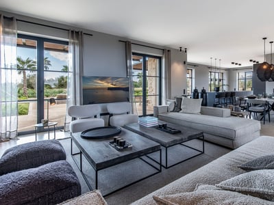 Salón moderno con sofás grises, mesas de madera y comedor con vistas al exterior a través de grandes ventanales.