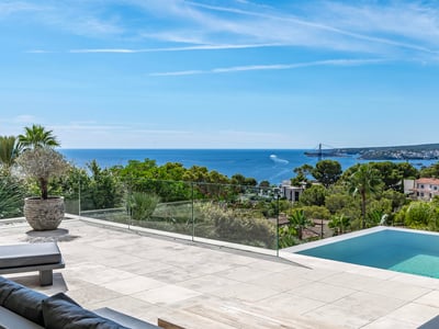 Terraza moderna de piedra con piscina infinita y barandilla de cristal con vistas al mar azul y vegetación exuberante.