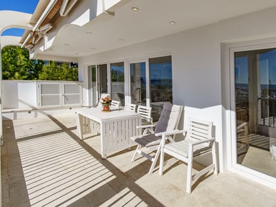 Terrasse ensoleillée avec table et chaises blanches, grandes baies vitrées reflétant la vue sur la côte.