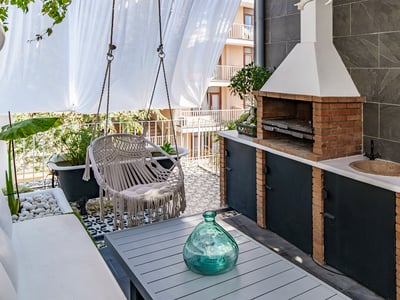 A modern terrace featuring a brick barbecue, a hanging macrame chair, a dining table, and decorative tiling.
