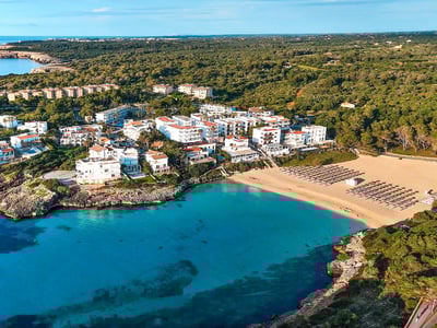 Drönarvy över en turkos vik med sandstrand omgiven av vita byggnader och lummig grön skog.