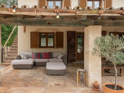 Covered terrace with wooden beams, stone pillars, comfortable sofa, and a potted olive tree.