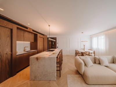 Modernt rum med öppen planlösning, köksö i marmor, träskåp, fiskbensparkett och stor beige soffa.