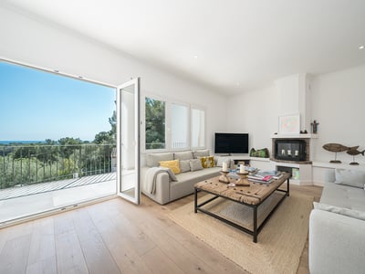 Luftigt vardagsrum med soffor, rustikt träbord, öppen spis och balkong med utsikt över träd och hav.