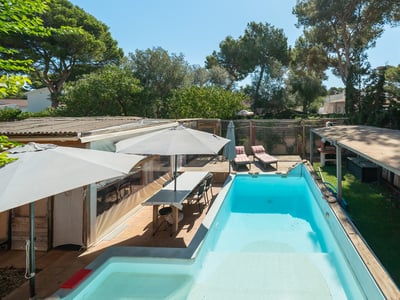 Piscina rectangular turquesa rodeada de tarima de madera, comedor exterior y árboles bajo un cielo azul despejado.