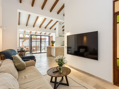 Modern living room featuring a beige sofa, wooden ceiling beams, large flat-screen TV, and a view into an open kitchen.