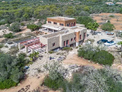 Vista aérea de una gran villa de piedra en construcción rodeada de densa vegetación mediterránea.