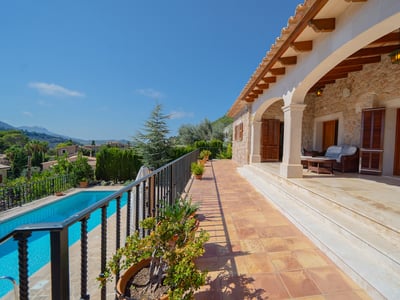 Terraza sombreada con vigas de madera, barandilla de hierro y vistas a una piscina azul y montañas lejanas.