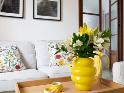 Sala de estar luminosa con sofá blanco, cojines florales y un jarrón amarillo vibrante sobre mesa de madera.