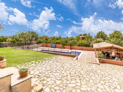 Gran patio de piedra con piscina, césped verde y una zona de estar cubierta con vigas de madera y techo de tejas.