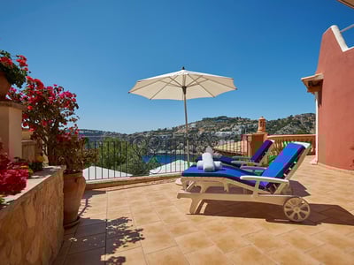 Terraza soleada con dos tumbonas azules, sombrilla blanca y vistas panorámicas a la costa y colinas.