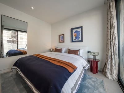 Dormitorio moderno con cama grande, ropa de cama azul y naranja, mesa roja y un gran espejo que refleja una ventana.