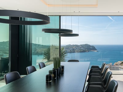 Modern black dining table and chairs facing floor-to-ceiling glass walls overlooking the blue ocean and coastline.