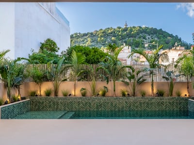 Modern pool med gröna fiskbensmönstrade kakelplattor omgiven av tropiska växter och utsikt över en kulle.