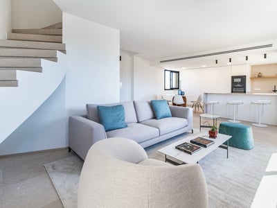 Salón moderno con sofá gris, cojines verde azulado, mesa de mármol y escalera de caracol junto a cocina blanca.