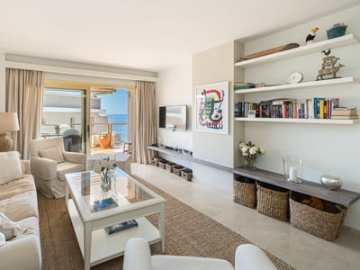 Salón de diseño mediterráneo con sofás beige, mesa de cristal y puertas correderas que dan a una terraza con mar al fondo.