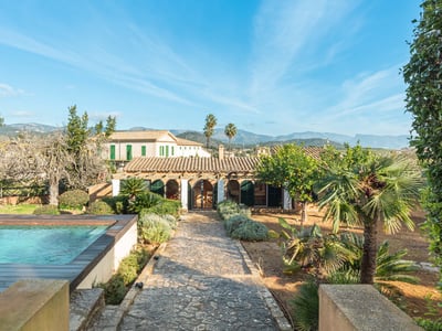 Piscina exterior y camino de piedra hacia una villa mediterránea con fondo de montañas bajo un cielo azul.