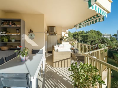 Amplio balcón con mesa de comedor exterior, sofá de mimbre y toldo de rayas verdes y blancas con vistas a los árboles.