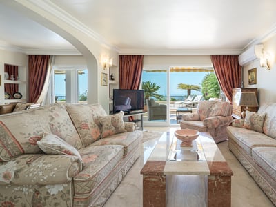 Spacious living room with floral sofas, marble table, and large glass doors opening to a terrace overlooking the sea.