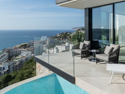 Modern infinity pool and terrace with grey armchairs overlooking the Mediterranean sea and a coastal town.