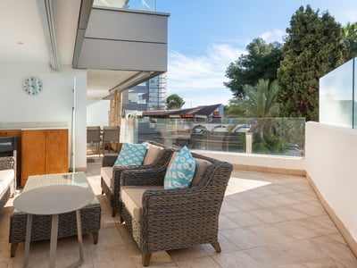 Rymlig terrass med grå rottingsoffor, blå kuddar och glasräcke under en klarblå himmel.